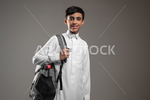 النظر إلى الكاميرا بإيماءات السرور والسعادة، متابعة الدروس والواجبات اليومية، المدارس التعليمية في السعودية, بورتريه مقرب لولد عربي سعودي خليجي مبتسم يرتدي الثوب التقليدي وحقيبة الظهر, بداية العام الدراسي الجديد, أهمية التعليم في بناء المستقبل, خلفية رمادية، استخدام حساس، محتوى حساس