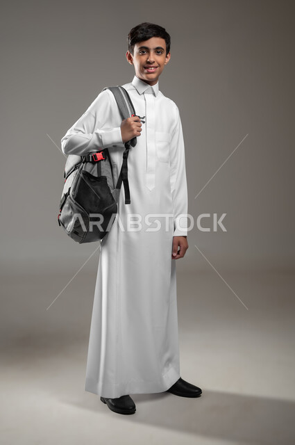 متابعة الدروس والواجبات اليومية، النظر إلى الكاميرا بإيماءات السرور والسعادة، المدارس التعليمية في السعودية, بورتريه لولد عربي سعودي خليجي مبتسم يرتدي الثوب التقليدي وحقيبة الظهر يستعد لبداية العام الدراسي الجديد, صورة الجسم بالكامل، خلفية رمادية، محتوى حساس، استخدام حساس