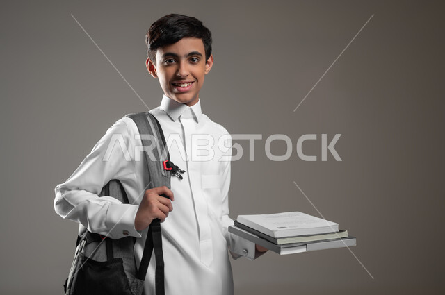 المدارس التعليمية في السعودية، متابعة الدروس والواجبات، النظر إلى الكاميرا بتعابير السرور, بورتريه مقرب لولد عربي سعودي خليجي مبتسم يرتدي الثوب التقليدي وحقيبة الظهر يمسك كتبه المدرسية، الاستعداد لبداية العام الدراسي الجديد, خلفية رمادية، محتوى حساس، استخدام حساس