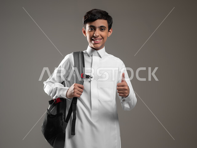 النظر إلى الكاميرا بتعابير القبول والرضا, بورتريه مقرب لولد عربي سعودي خليجي مبتسم يرتدي الثوب التقليدي وحقيبة الظهر يستعد لبداية العام الدراسي الجديد, المدارس التعليمية في السعودية، متابعة الدروس والواجبات، خلفية رمادية، استخدام حساس، محتوى حساس