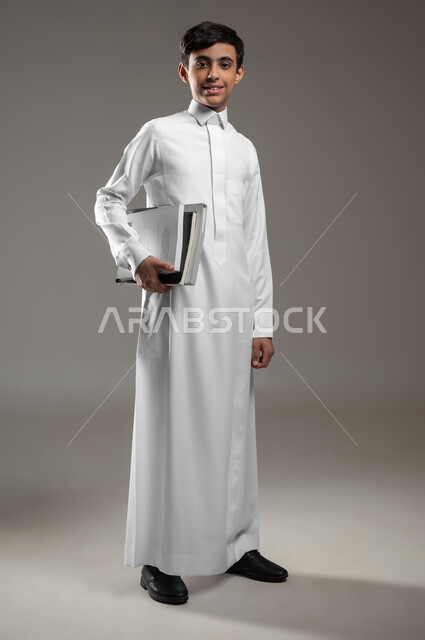 الوقوف باستقامة والنظر إلى الكاميرا بتعابير السرور, بورتريه لولد عربي سعودي خليجي مبتسم يرتدي الثوب التقليدي ويمسك كتبه المدرسية، الاستعداد لبداية العام الدراسي, المدارس التعليمية في السعودية، متابعة الدروس والواجبات، صورة الجسم بالكامل، خلفية رمادية، محتوى حساس، استخدام حساس