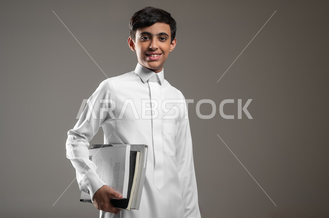 الوقوف باستقامة والنظر إلى الكاميرا بتعابير السرور, بورتريه مقرب لولد عربي سعودي خليجي مبتسم يرتدي الثوب التقليدي ويمسك بيده كتبه المدرسية، الاستعداد لبداية العام الدراسي الجديد, المدارس التعليمية في السعودية، متابعة الدروس والواجبات، خلفية رمادية، محتوى حساس، استخدام حساس