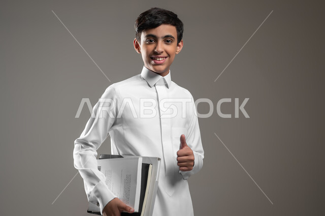 النظر إلى الكاميرا بتعابير القبول والرضا والإعجاب, بورتريه مقرب لولد عربي سعودي خليجي مبتسم يرتدي الثوب التقليدي يمسك كتبه المدرسية، الاستعداد لبداية العام الدراسي الجديد, المدارس التعليمية في السعودية، متابعة الدروس والواجبات، خلفية رمادية، محتوى حساس، استخدام حساس