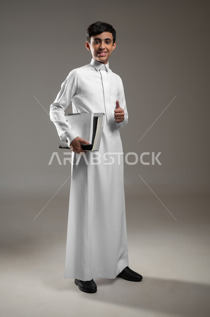 الاستعداد لبداية العام الدراسي الجديد, النظر إلى الكاميرا بتعابير القبول والرضا والإعجاب, بورتريه لولد عربي سعودي خليجي مبتسم يرتدي الثوب التقليدي يمسك كتبه المدرسية، المدارس التعليمية في السعودية، متابعة الدروس والواجبات، صورة الجسم بالكامل، خلفية رمادية، محتوى حساس، استخدام حساس