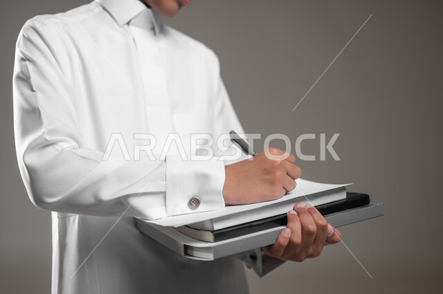 الاجتهاد وتحضير الدروس, حل وكتابة الواجبات اليومية، بداية العام الدراسي الجديد, تطوير المناهج التعليمية في السعودية, بورتريه مقرب لولد عربي سعودي خليجي مبتسم يرتدي الثوب التقليدي يقوم بتسجيل وتدوين الملاحظات, خلفية رمادية، محتوى حساس، استخدام حساس