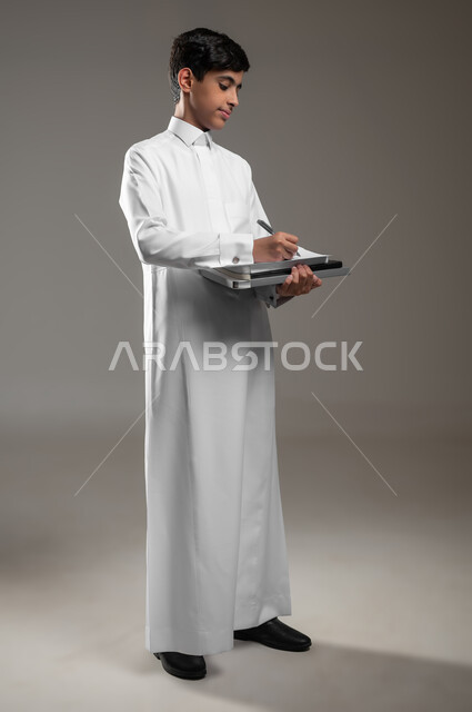 Recording and taking notes, diligence and preparing lessons, the beginning of the new school year, developing educational curricula in Saudi Arabia, a portrait of a smiling Saudi Arabian Gulf boy wearing a traditional dress solving and writing daily assignments, full-length body image, gray background, sensitive content, sensitive use