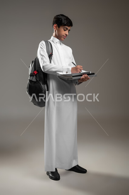 تسجيل وتدوين الملاحظات، الاجتهاد وتحضير الدروس, بداية العام الدراسي الجديد, تطوير المناهج التعليمية في السعودية, بورتريه لولد عربي سعودي خليجي مبتسم يرتدي الثوب التقليدي يحمل حقيبة الظهر يقوم بحل وكتابة الواجبات اليومية, صورة الجسم بالطول الكامل، خلفية رمادية، محتوى حساس، استخدام حساس
