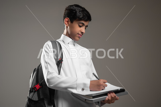 حل وكتابة الواجبات اليومية، بداية العام الدراسي الجديد, الاجتهاد وتحضير الدروس, تطوير المناهج التعليمية في السعودية, بورتريه مقرب لولد عربي سعودي خليجي مبتسم يرتدي الثوب التقليدي يحمل الحقيبة المدرسية يقوم بتسجيل وتدوين الملاحظات, خلفية رمادية، محتوى حساس، استخدام حساس