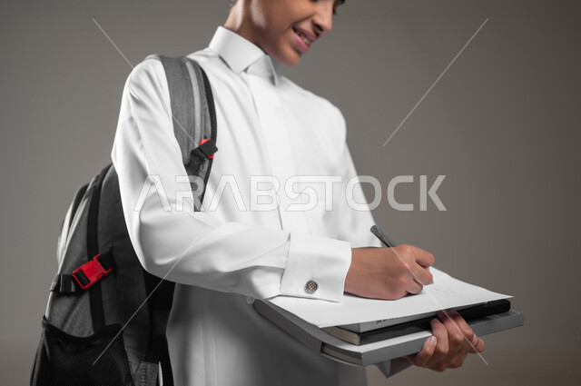 حل وكتابة الواجبات اليومية، بداية العام الدراسي الجديد, الاجتهاد وتحضير الدروس, تطوير المناهج التعليمية في السعودية, بورتريه مقرب لولد عربي سعودي خليجي مبتسم يرتدي الثوب التقليدي يحمل الحقيبة المدرسية يقوم بتسجيل وتدوين الملاحظات, خلفية رمادية، محتوى حساس، استخدام حساس