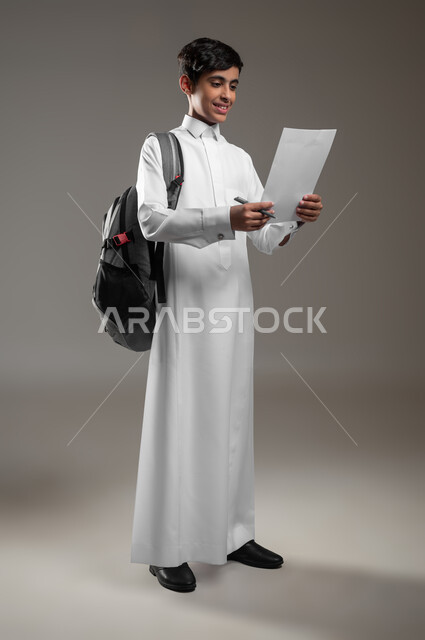 تطوير المناهج التعليمية في السعودية, متابعة الواجبات اليومية، الاجتهاد وتحضير الدروس، بورتريه لولد عربي سعودي خليجي مبتسم يرتدي الثوب التقليدي يحمل الحقيبة المدرسية يقوم بقراءة الأوراق, بداية العام الدراسي الجديد, صورة الجسم بالكامل، خلفية رمادية، محتوى حساس، استخدام حساس
