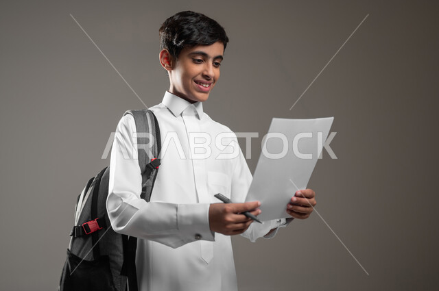 حل الواجبات اليومية، الاجتهاد وتحضير الدروس، تطوير المناهج التعليمية في السعودية, بورتريه مقرب لولد عربي سعودي خليجي مبتسم يرتدي الثوب التقليدي يحمل الحقيبة المدرسية يقوم بقراءة الأوراق, بداية العام الدراسي الجديد, خلفية رمادية، محتوى حساس، استخدام حساس