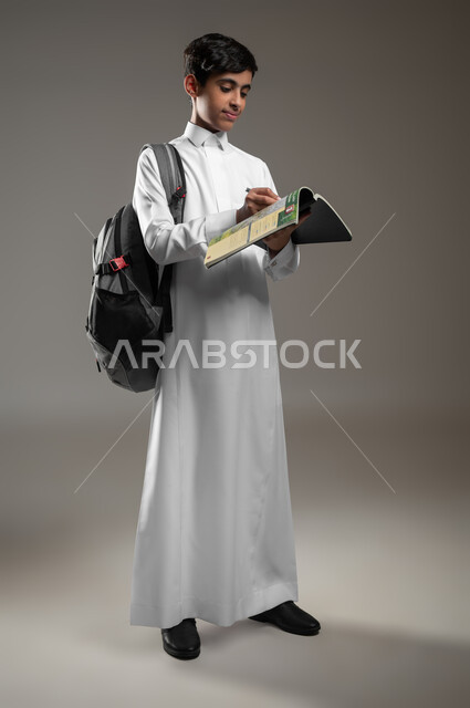تطوير المناهج التعليمية في السعودية, الاجتهاد وتحضير الدروس, متابعة الواجبات اليومية، بورتريه لولد عربي سعودي خليجي مبتسم يرتدي الثوب التقليدي يحمل الحقيبة المدرسية يقوم بقراءة الكتب, صورة الجسم بالكامل، خلفية رمادية، محتوى حساس، استخدام حساس