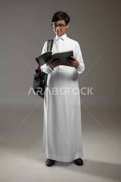تطور المناهج التعليمية في السعودية, بورتريه لولد عربي سعودي خليجي يرتدي الثوب التقليدي يضع النظارات الطبية يحمل الحقيبة المدرسية يقوم بقراءة الكتب, بداية العام الدراسي الجديد, الاجتهاد وتحضير الدروس, صورة الجسم بالكامل، خلفية رمادية، محتوى حساس، استخدام حساس