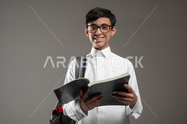 الاجتهاد وتحضير الدروس, تطور المناهج التعليمية في المدارس السعودية, بورتريه مقرب لولد عربي سعودي خليجي مبتسم يرتدي الثوب التقليدي يضع النظارات الطبية يحمل الحقيبة المدرسية يقوم بقراءة الكتب, بداية العام الدراسي الجديد, خلفية رمادية، محتوى حساس، استخدام حساس