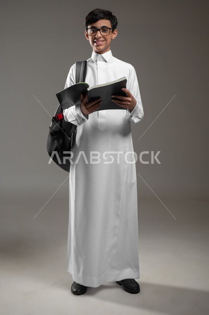 تطور المناهج التعليمية في السعودية, بورتريه لولد عربي سعودي خليجي يرتدي الثوب التقليدي والنظارات الطبية يحمل الحقيبة المدرسية يقوم بقراءة الكتب, بداية العام الدراسي الجديد, الاجتهاد وتحضير الدروس, صورة الجسم بالكامل، خلفية رمادية، محتوى حساس، استخدام حساس
