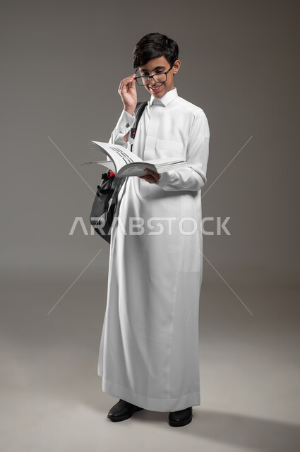 متابعة الدروس والواجبات، الاجتهاد وتحضير الدروس, تطوير المناهج التعليمية في السعودية, بورتريه لولد عربي سعودي خليجي يرتدي الثوب التقليدي يضع النظارات الطبية يحمل الحقيبة المدرسية يقوم بقراءة الكتب, صورة الجسم بالكامل، خلفية رمادية، محتوى حساس، استخدام حساس