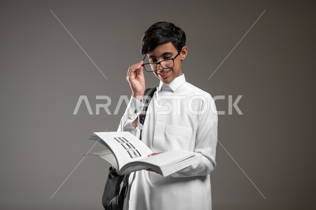 الاجتهاد وتحضير الدروس, تطوير المناهج التعليمية في السعودية, بورتريه مقرب لولد عربي سعودي خليجي مبتسم يرتدي الثوب التقليدي يضع النظارات الطبية يحمل الحقيبة المدرسية يقوم بقراءة الكتب, بداية العام الدراسي الجديد, خلفية رمادية، محتوى حساس، استخدام حساس