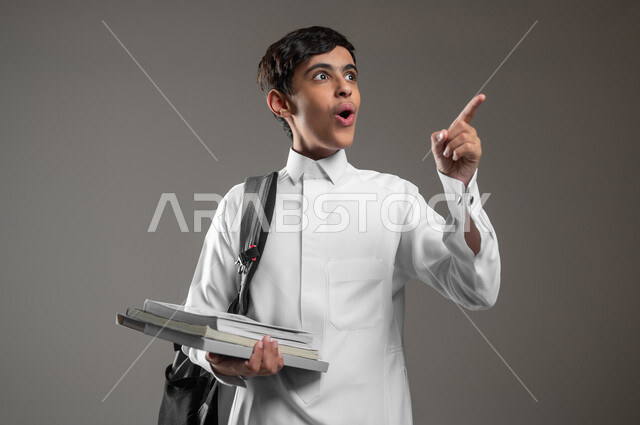 الإشارة الى شيء ما بتعابير الاستغراب والدهشة، متابعة الدروس والواجبات, بورتريه مقرب لولد عربي سعودي خليجي متفاجئ يرتدي الثوب التقليدي وحقيبة الظهر يمسك بيده كتبه المدرسية، الاستعداد لبداية العام الدراسي الجديد, خلفية رمادية، محتوى حساس، استخدام حساس