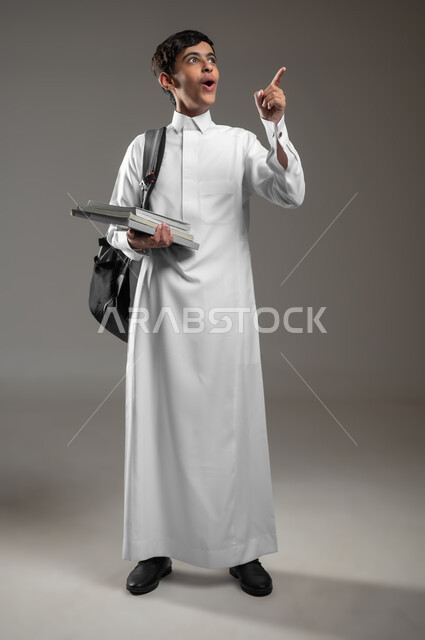 الإشارة الى شيء ما بتعابير الاستغراب والدهشة، متابعة الدروس والواجبات, بورتريه لولد عربي سعودي خليجي متفاجئ يرتدي الثوب التقليدي وحقيبة الظهر يمسك كتبه المدرسية، الاستعداد لبداية العام الدراسي الجديد, صورة الجسم بالكامل، خلفية رمادية، محتوى حساس، استخدام حساس