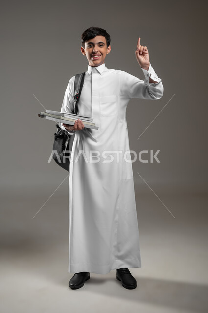 المشاركة في الحصص والإجابة على الاسئلة، متابعة الدروس والواجبات, بورتريه لولد عربي سعودي خليجي مبتسم يرتدي الثوب التقليدي وحقيبة الظهر يمسك كتبه المدرسية يرفع اصبعه للاعلى بتعابير السرور، صورة الجسم بالكامل، خلفية رمادية، محتوى حساس، استخدام حساس