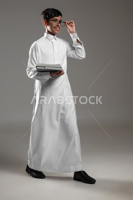 المدارس التعليمية في المملكة السعودية، بورتريه لولد عربي سعودي خليجي مبتسم يرتدي الثوب التقليدي والنظارات الطبية يمسك كتبه المدرسية ينظر الى شيء ما، الاستعداد لبداية العام الدراسي الجديد, صورة الجسم بالكامل، خلفية رمادية، محتوى حساس، استخدام حساس