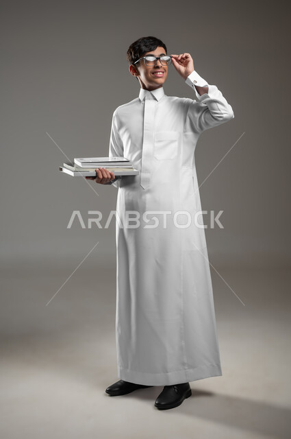 المدارس التعليمية في المملكة السعودية، بورتريه لولد عربي سعودي خليجي مبتسم يرتدي الثوب التقليدي والنظارات الطبية يمسك كتبه المدرسية ينظر الى شيء ما، الاستعداد لبداية العام الدراسي الجديد, صورة الجسم بالكامل، خلفية رمادية، محتوى حساس، استخدام حساس