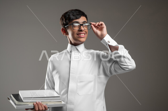 النظر الى شيء ما بإيماءات السرور، المدارس التعليمية في المملكة السعودية، بورتريه مقرب لولد عربي سعودي خليجي مبتسم يرتدي الثوب التقليدي والنظارات الطبية يمسك كتبه المدرسية، الاستعداد لبداية العام الدراسي الجديد, خلفية رمادية، محتوى حساس، استخدام حساس