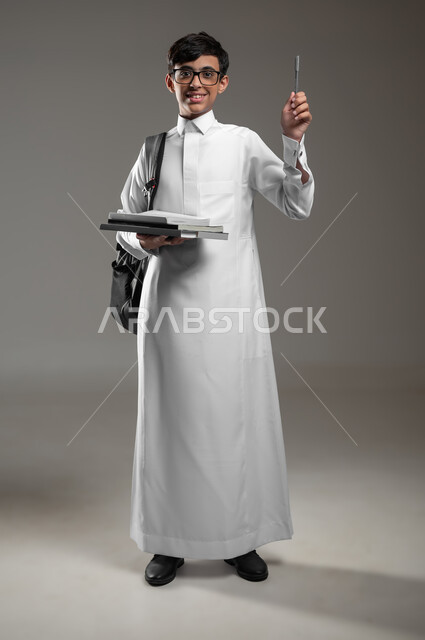المشاركة في الحصص والإجابة على الاسئلة، بورتريه لولد عربي سعودي خليجي مبتسم يرتدي الثوب التقليدي وحقيبة الظهر يمسك كتبه المدرسية يرفع قلمه للاعلى بتعابير السرور، الاستعداد لبداية العام الدراسي الجديد, صورة الجسم بالكامل، خلفية رمادية، محتوى حساس، استخدام حساس
