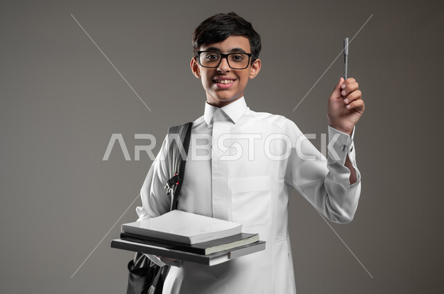 متابعة الدروس والواجبات, المشاركة في الحصص والإجابة على الاسئلة، بورتريه مقرب لولد عربي سعودي خليجي يرتدي الثوب التقليدي وحقيبة الظهر يمسك كتبه المدرسية يرفع قلمه للاعلى بتعابير السرور، الاستعداد لبداية العام الدراسي، خلفية رمادية، محتوى حساس، استخدام حساس