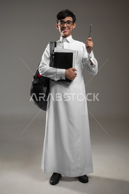 المشاركة في الحصص والإجابة على الاسئلة، بورتريه لولد عربي سعودي خليجي مبتسم يرتدي الثوب التقليدي وحقيبة الظهر يمسك كتبه المدرسية يرفع قلمه للاعلى بتعابير السرور، الاستعداد لبداية العام الدراسي الجديد, صورة الجسم بالكامل، خلفية رمادية، محتوى حساس، استخدام حساس