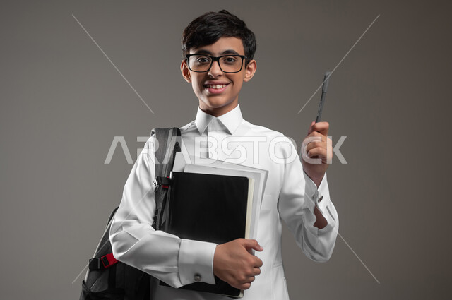 متابعة الدروس والواجبات, المشاركة في الحصص والإجابة على الاسئلة، بورتريه مقرب لولد عربي سعودي خليجي مبتسم يرتدي الثوب التقليدي وحقيبة الظهر يمسك كتبه ويرفع قلمه للاعلى بتعابير السرور، الاستعداد لبداية العام الدراسي, خلفية رمادية، محتوى حساس، استخدام حساس