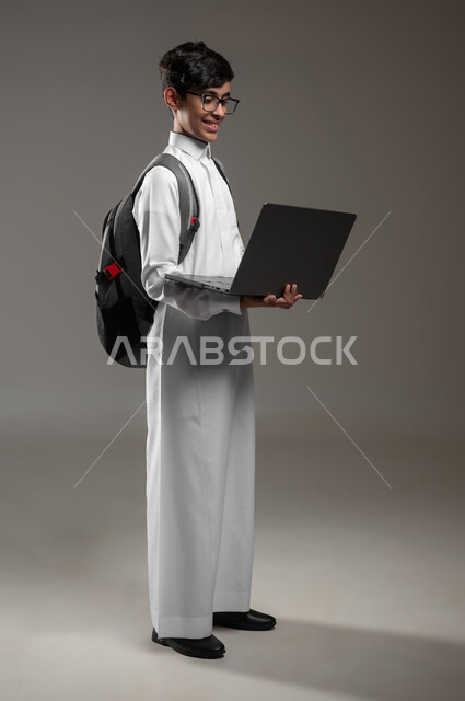 ادخال التقية والتكنولوجيا بالتعليم، متابعة منصات التعليم الالكتروني بإستعمال جهاز الحاسب المحمول، بورتريه لولد عربي خليجي سعودي يرتدي الثوب التقليدي وحقيبة الظهر يقوم بحل الواجبات المنزليه اونلاين، صورة الجسم بالكامل، خلفية رمادية، محتوى حساس، استخدام حساس
