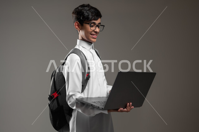 ادخال التقنية والتكنولوجيا بالتعليم، متابعة منصات التعليم الالكتروني عبر الانترنت بإستعمال جهاز الحاسب المحمول، بورتريه مقرب لولد عربي خليجي سعودي يرتدي الثوب التقليدي وحقيبة الظهر يقوم بحل الواجبات المنزليه عبر اللابتوب، خلفية رمادية، محتوى حساس، استخدام حساس