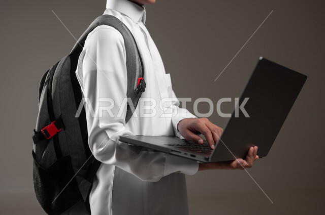 دمج التقنية والتكنولوجيا بالتعليم، متابعة منصات التعليم الالكتروني عبر الانترنت بإستخدام جهاز الحاسب المحمول، بورتريه مقرب لولد عربي خليجي سعودي يرتدي الثوب التقليدي وحقيبة الظهر يقوم بحل الواجبات المنزليه عبر اللابتوب، خلفية رمادية، محتوى حساس، استخدام حساس