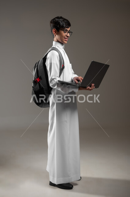 Following e-learning platforms using a laptop, integrating technology and technology into education, portrait of a Saudi Gulf Arab boy wearing a traditional dress and a backpack doing homework via the laptop, full body image, gray background, sensitive content, sensitive use