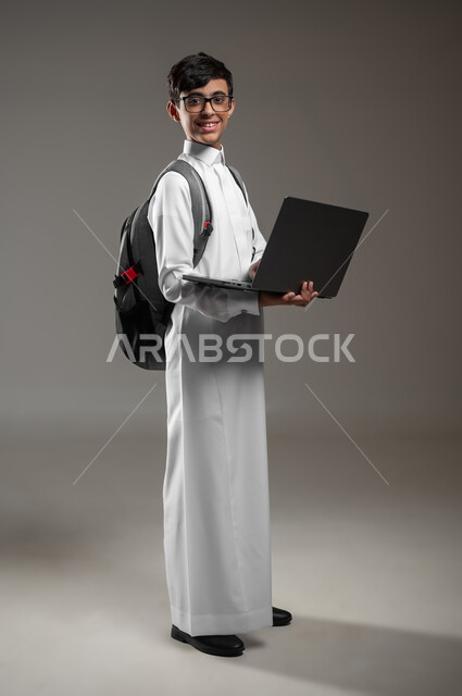 متابعة منصات التعليم الالكتروني باستعمال جهاز الحاسب المحمول، دمج التقنية والتكنولوجيا بالتعليم، بورتريه لولد عربي خليجي سعودي يرتدي الثوب التقليدي وحقيبة الظهر يقوم بحل الواجبات المنزليه عبر اللابتوب، صورة الجسم بالكامل، خلفية رمادية، محتوى حساس، استخدام حساس