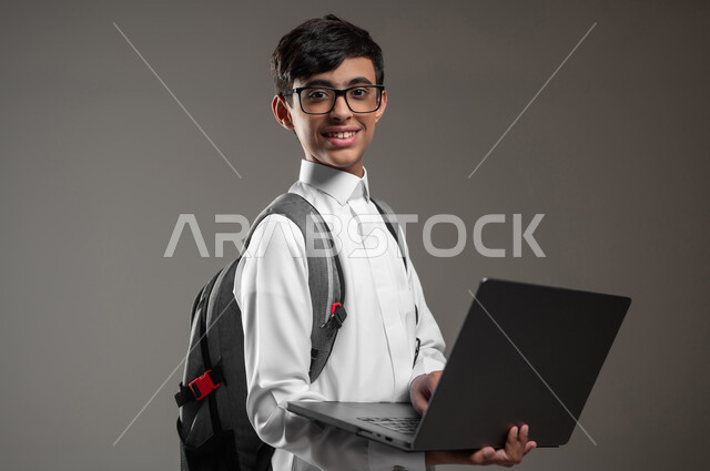 دمج التقنية والتكنولوجيا بالتعليم، متابعة منصات التعليم الالكتروني عبر الانترنت بإستعمال جهاز الحاسب المحمول، بورتريه مقرب لولد عربي خليجي سعودي مبتسم يرتدي الثوب التقليدي وحقيبة الظهر يقوم بحل الواجبات المنزليه اونلاين، خلفية رمادية، استخدام حساس، محتوى حساس