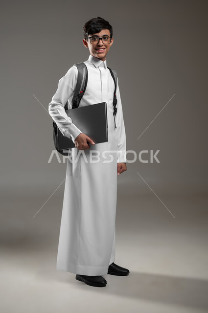 The use of modern technical devices in the scientific field, a portrait of a smiling Saudi Arabian Gulf boy wearing a traditional dress and a backpack holding a laptop in his hand, preparing for the beginning of the school year, full body portrait, gray background, sensitive use, sensitive content