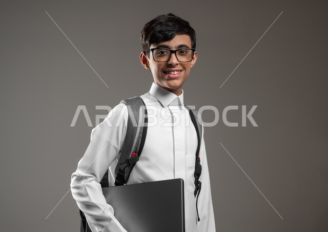 استخدام الأجهزة التقنية الحديثة في المجال العلمي، دمج التقنية والتكنولوجيا بالتعليم، بورتريه مقرب لولد عربي خليجي سعودي مبتسم يرتدي الثوب التقليدي وحقيبة الظهر يحمل بيده الحاسوب المحمول، الاستعداد لبداية العام الدراسي، خلفية رمادية، محتوى حساس، استخدام حساس