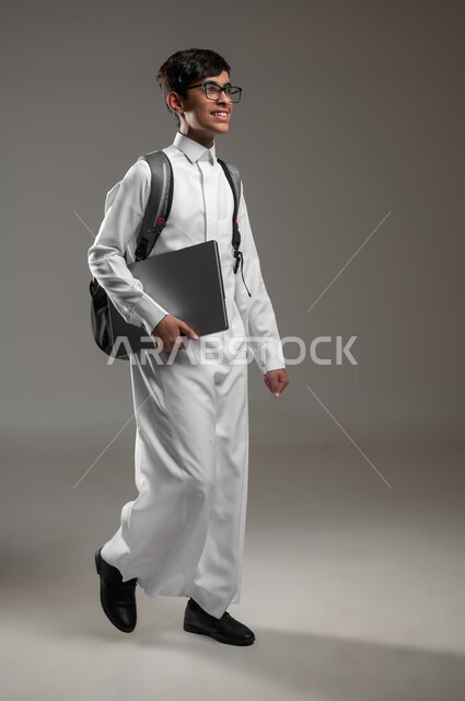 ايماءات المشي والذهاب الى المدرسة، دمج التقنية والتكنولوجيا الحديثة بالتعليم، بورتريه مقرب لولد عربي خليجي سعودي مبتسم يرتدي الثوب التقليدي وحقيبة الظهر يحمل بيده جهاز الحاسب المحمول، صورة الجسم بالكامل، خلفية رمادية، محتوى حساس، استخدام حساس