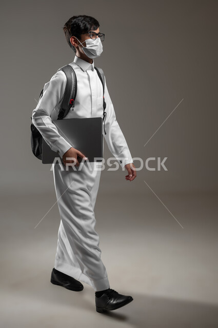 ارتداء الكمامة للوقاية من الأمراض، متابعة الدروس والواجبات، تعابير وإيماءات المشي والذهاب إلى المدرسة، بورتريه لولد عربي سعودي خليجي يرتدي الثوب التقليدي وحقيبة الظهر يستعد لبداية العام الدراسي الجديد, صورة الجسم بالكامل، خلفية رمادية، محتوى حساس، استخدام حساس