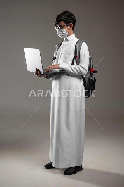 دمج التقنية والتكنولوجيا بالتعليم، ارتداء الكمامة للوقاية من الأمراض، بورتريه لولد عربي خليجي سعودي يرتدي الثوب التقليدي وحقيبة الظهر يقوم بمتابعة منصات التعليم الالكتروني بإستعمال الحاسب المحمول، صورة الجسم بالكامل، خلفية رمادية، محتوى حساس، استخدام حساس
