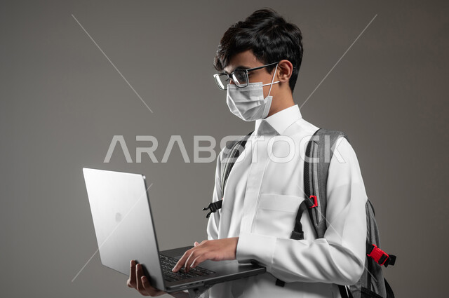 ارتداء الكمامة للوقاية من الأمراض، دمج التقنية والتكنولوجيا بالتعليم، بورتريه لولد عربي خليجي سعودي يرتدي الثوب التقليدي وحقيبة الظهر يقوم بمتابعة منصات التعلم الالكتروني باستعمال الحاسوب المحمول، صورة الجسم بالكامل، خلفية رمادية، محتوى حساس، استخدام حساس