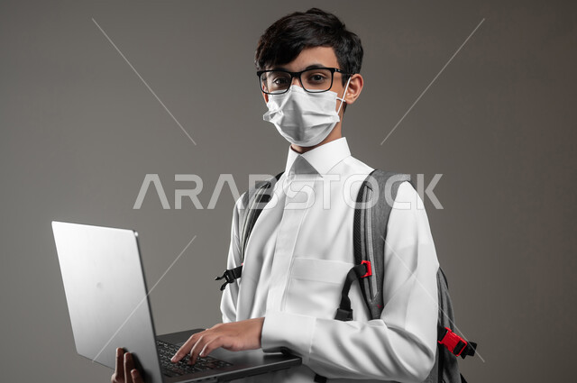 حل الواجبات بإستعمال اللابتوب، ارتداء الكمامة للوقاية من الأمراض، دمج التقنية والتكنولوجيا بالتعليم، بورتريه لولد عربي خليجي سعودي يرتدي الثوب التقليدي وحقيبة الظهر يقوم بمتابعة منصات التعلم الالكتروني، صورة لكامل الجسم، خلفية رمادية، محتوى حساس، استخدام حساس