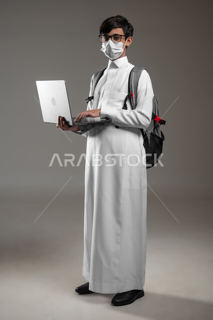 ارتداء الكمامة للوقاية من الأمراض، متابعة منصات التعلم الالكتروني بإستعمال اللابتوب، دمج التكنولوجيا بالتعليم، بورتريه لولد عربي خليجي سعودي يرتدي الثوب التقليدي وحقيبة الظهر يقوم بحل الواجبات اونلاين، صورة الجسم بالكامل، خلفية رمادية، محتوى حساس، استخدام حساس