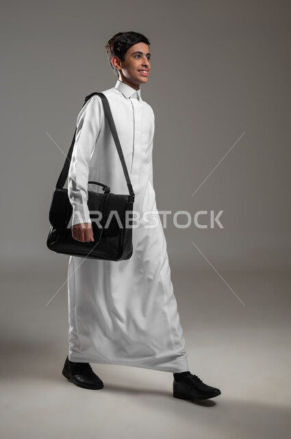 Expressions and gestures of walking and going to school, a portrait of a smiling Saudi Arabian Gulf boy wearing a traditional dress holding a leather bag preparing for the start of the new school year, following lessons and daily duties, full body image, gray background, sensitive content, sensitive use
