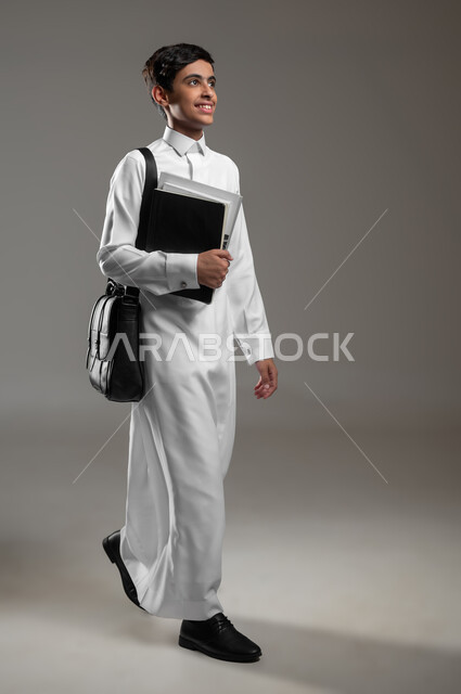 الاستعداد لبداية العام الدراسي الجديد, متابعة الدروس والواجبات اليومية، إيماءات المشي والذهاب الى المدرسة، بورتريه لولد عربي سعودي خليجي مبتسم يرتدي الثوب التقليدي وحقيبة جلد يمسك بيده الكتب، صورة الجسم بالكامل، خلفية رمادية، محتوى حساس، استخدام حساس