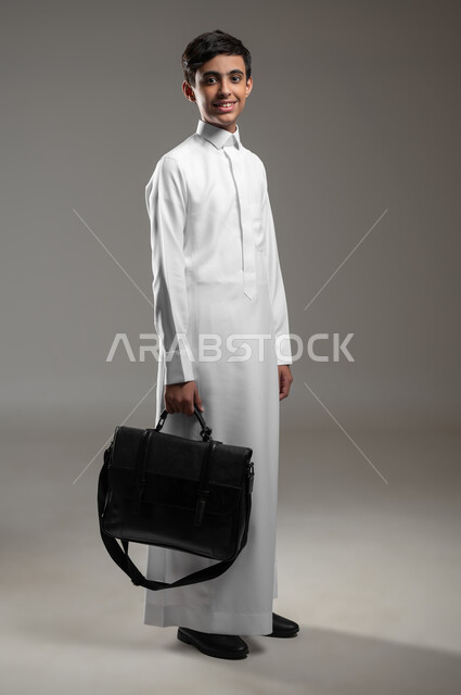النظر إلى الكاميرا بإيماءات السرور والسعادة، المدارس التعليمية في السعودية, بورتريه لولد عربي سعودي خليجي مبتسم يرتدي الثوب التقليدي يمسك حقيبة جلد يستعد لبداية العام الدراسي الجديد, صورة الجسم بالكامل، خلفية رمادية، محتوى حساس، استخدام حساس