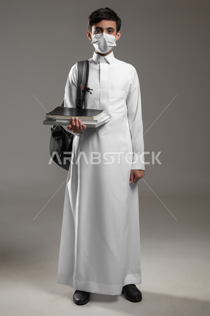الاستعداد لبداية العام الدراسي الجديد, ارتداء الكمامة للوقاية من الأمراض والعدوى، متابعة الدروس والواجبات, بورتريه لولد عربي سعودي خليجي يرتدي الثوب التقليدي وحقيبة الظهر يمسك كتبه المدرسية، صورة الجسم بالكامل، خلفية رمادية، محتوى حساس، استخدام حساس