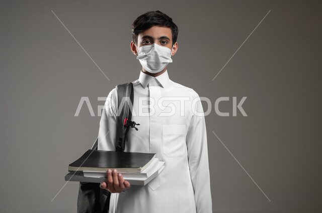 ارتداء الكمامة للوقاية من الأمراض، المدارس التعليمية في السعودية، متابعة الدروس والواجبات, بورتريه مقرب لولد عربي سعودي خليجي يرتدي الثوب التقليدي وحقيبة الظهر يمسك كتبه المدرسية، الاستعداد لبداية العام الدراسي الجديد, خلفية رمادية، محتوى حساس، استخدام حساس